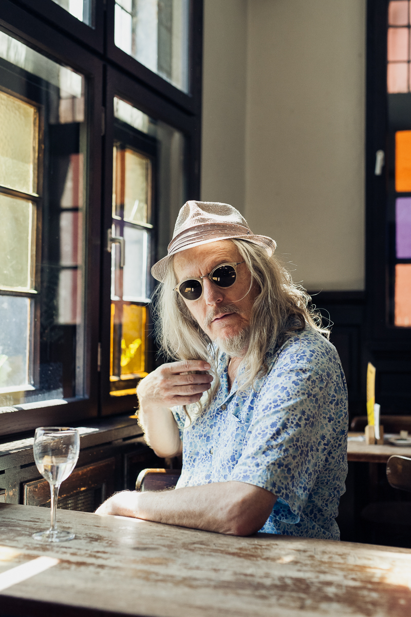 An einem Tisch am Fenster sitzt eine Person mit langen Haaren und Sonnenbrille. Sie trägt ein gemustertes Hemd und einen Hut, der an den einzigartigen Stil Wolfgang Beltracchis erinnert, und davor steht ein leeres Weinglas. Portrait und Reportage Fotografie für Corporate und Editorial