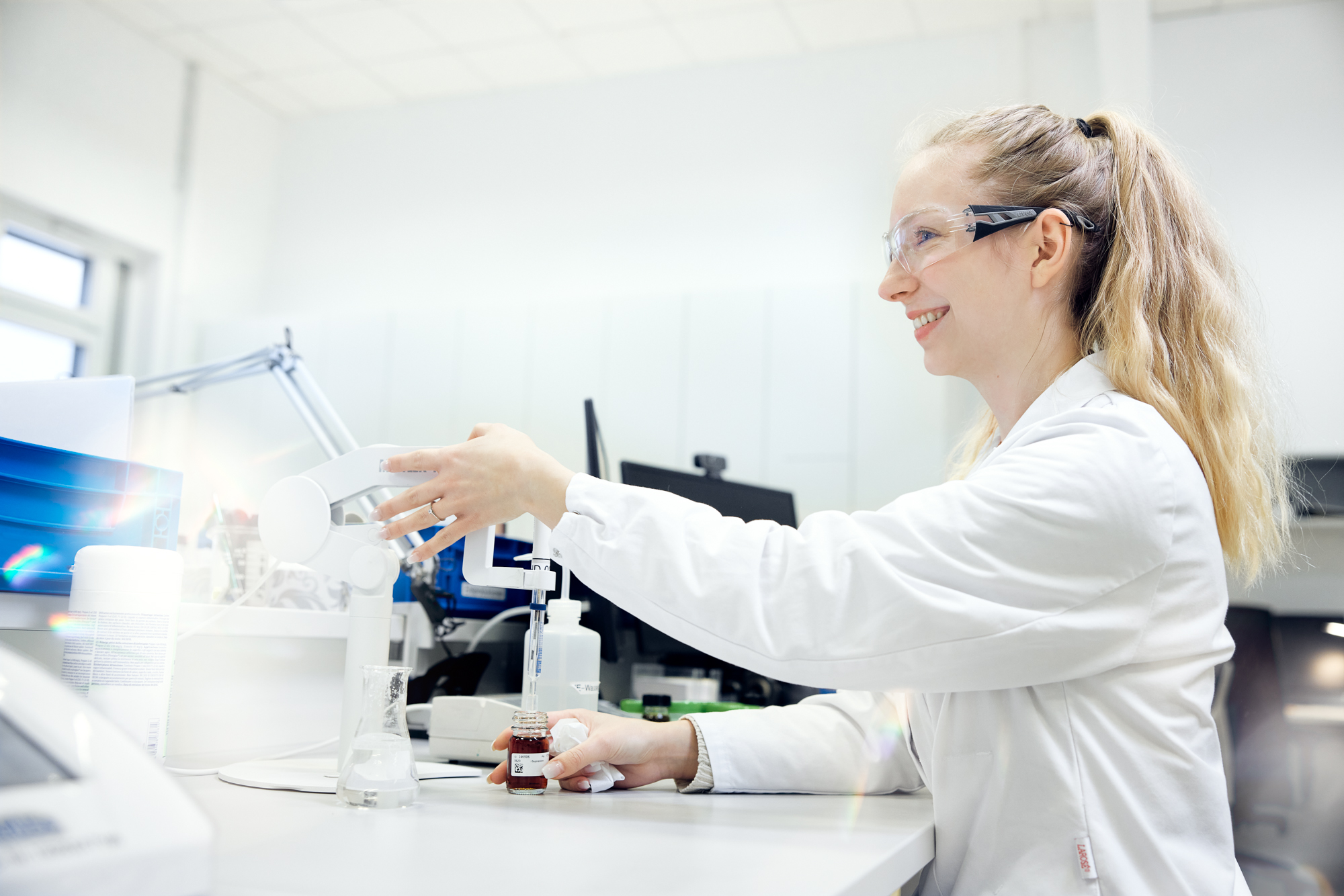 Ein Wissenschaftler im Laborkittel und mit Schutzbrille führt ein Experiment durch, bei dem er Flüssigkeit aus einer Flasche in einen Becher gießt. Diese Szene fängt die Essenz der Unternehmensfotografie in Düsseldorf ein, umgeben von Laborgeräten im hellen Labor. Portrait und Reportage Fotografie für Corporate und Editorial