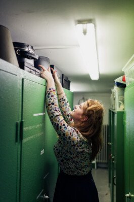 In einem Raum mit grünen Schließfächern und Neonlicht zeigt ein Unternehmensporträt jemanden, der auf einem hohen Regal nach einem Hut greift – ein faszinierender Moment im Düsseldorfer Geschäftsalltag. Portrait und Reportage Fotografie für Corporate und Editorial