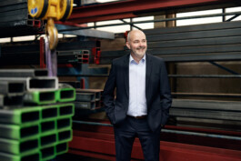 Ein Mann im Anzug steht mit den Händen in den Taschen in einer Lagerhalle, umgeben von gestapelten Metallträgern – eine ideale Kulisse für die Unternehmensfotografie in Düsseldorf, die die Essenz von Industrie und Professionalität einfängt. Portrait und Reportage Fotografie für Corporate und Editorial