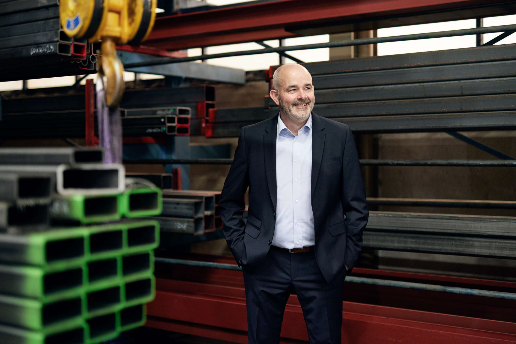 Ein Mann im Anzug steht mit den Händen in den Taschen in einer Lagerhalle, umgeben von gestapelten Metallträgern – eine ideale Kulisse für die Unternehmensfotografie in Düsseldorf, die die Essenz von Industrie und Professionalität einfängt. Portrait und Reportage Fotografie für Corporate und Editorial
