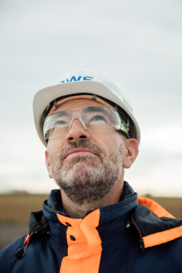 Ein Mann mit Schutzhelm, Schutzbrille und Warnweste blickt in einen wolkigen Himmel und verkörpert die Essenz der Unternehmensfotografie in Düsseldorf. Portrait und Reportage Fotografie für Corporate und Editorial