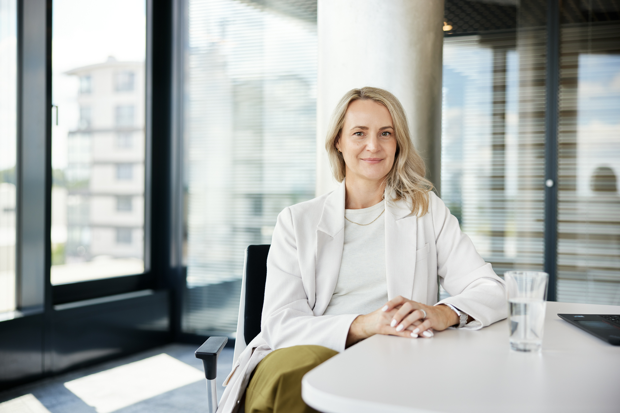 Eine Person sitzt an einem Tisch in einem modernen Büro in Düsseldorf, durch das große Fenster natürliches Licht hereinlassen. In einem hellen Outfit gekleidet und mit einem Glas Wasser in der Hand fängt diese Szene die Essenz der Unternehmensfotografie für Portraits und Reportagen für Unternehmen ein. Portrait und Reportage Fotografie für Corporate und Editorial