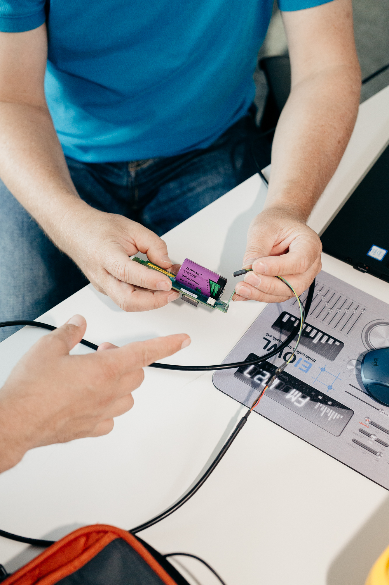 Zwei Personen arbeiten gemeinsam an einem kleinen elektronischen Gerät, dessen Kabel miteinander verbunden sind. Einer hält es, während der andere darauf zeigt. Auf einem Schreibtisch stehen eine Computermaus und ein Mauspad. So wird die Essenz der Teamarbeit in der Unternehmensfotografie für Düsseldorfer Unternehmen eingefangen. Portrait und Reportage Fotografie für Corporate und Editorial
