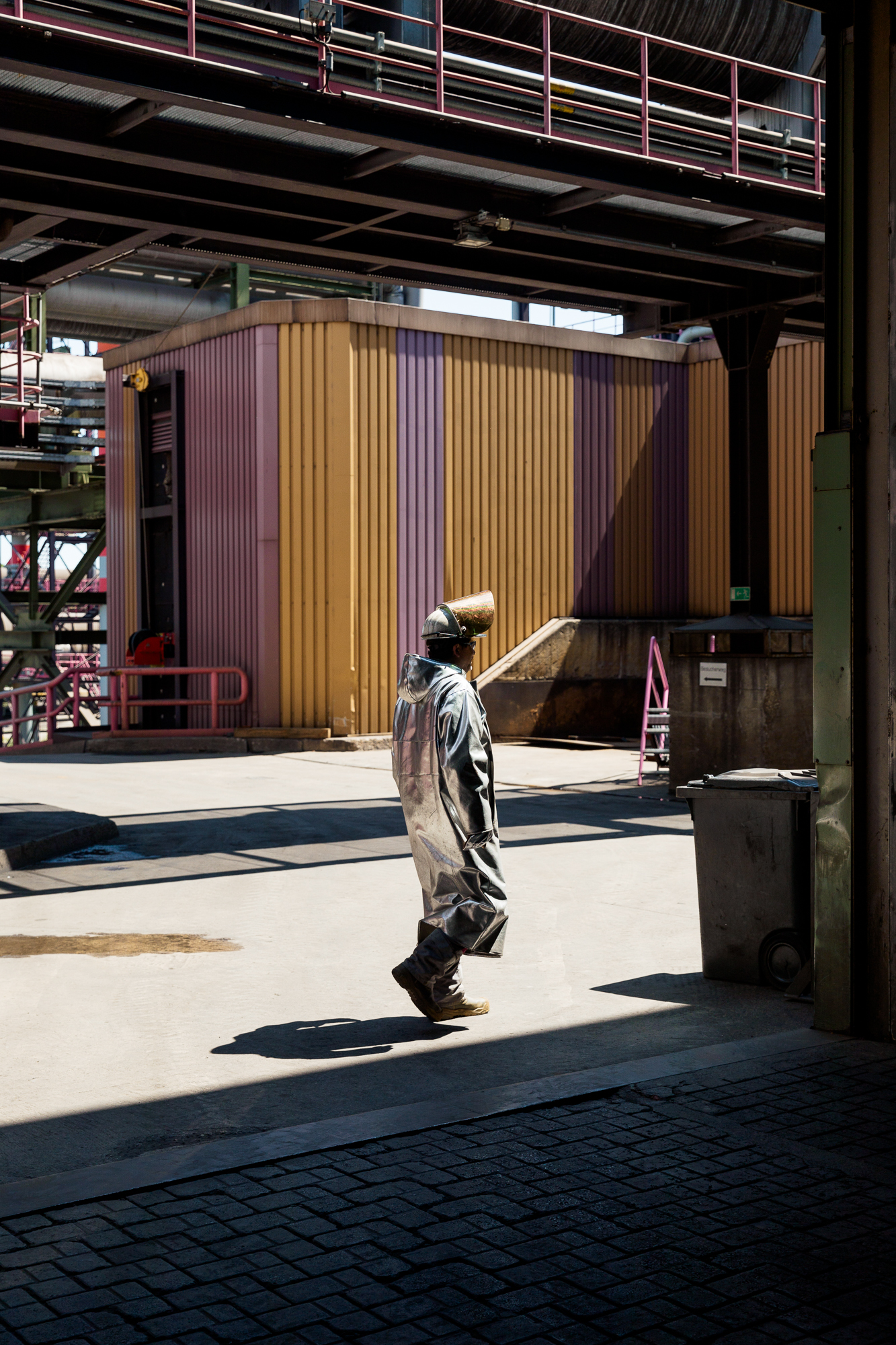 Eine Person in Schutzausrüstung navigiert durch ein Industriegelände mit Metallkonstruktionen und großen Gebäuden und fängt die Essenz der Unternehmensfotografie Düsseldorf durch eindrucksvolle Porträts und Reportagen für Unternehmen ein. Portrait und Reportage Fotografie für Corporate und Editorial