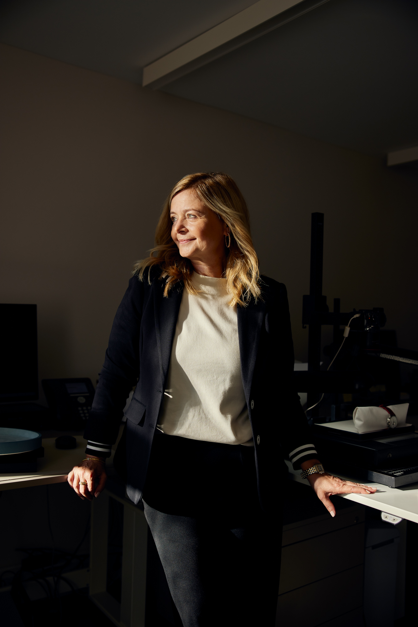 Eine Frau mit langen Haaren, Blazer und Pullover bekleidet, steht lässig in einem Büro und lehnt an einem Schreibtisch. Diese Szene fängt die Essenz von Corporate Fotografie Düsseldorf ein und verbindet gekonnt Professionalität und Zugänglichkeit für maßgeschneiderte Unternehmensporträts. Portrait und Reportage Fotografie für Corporate und Editorial