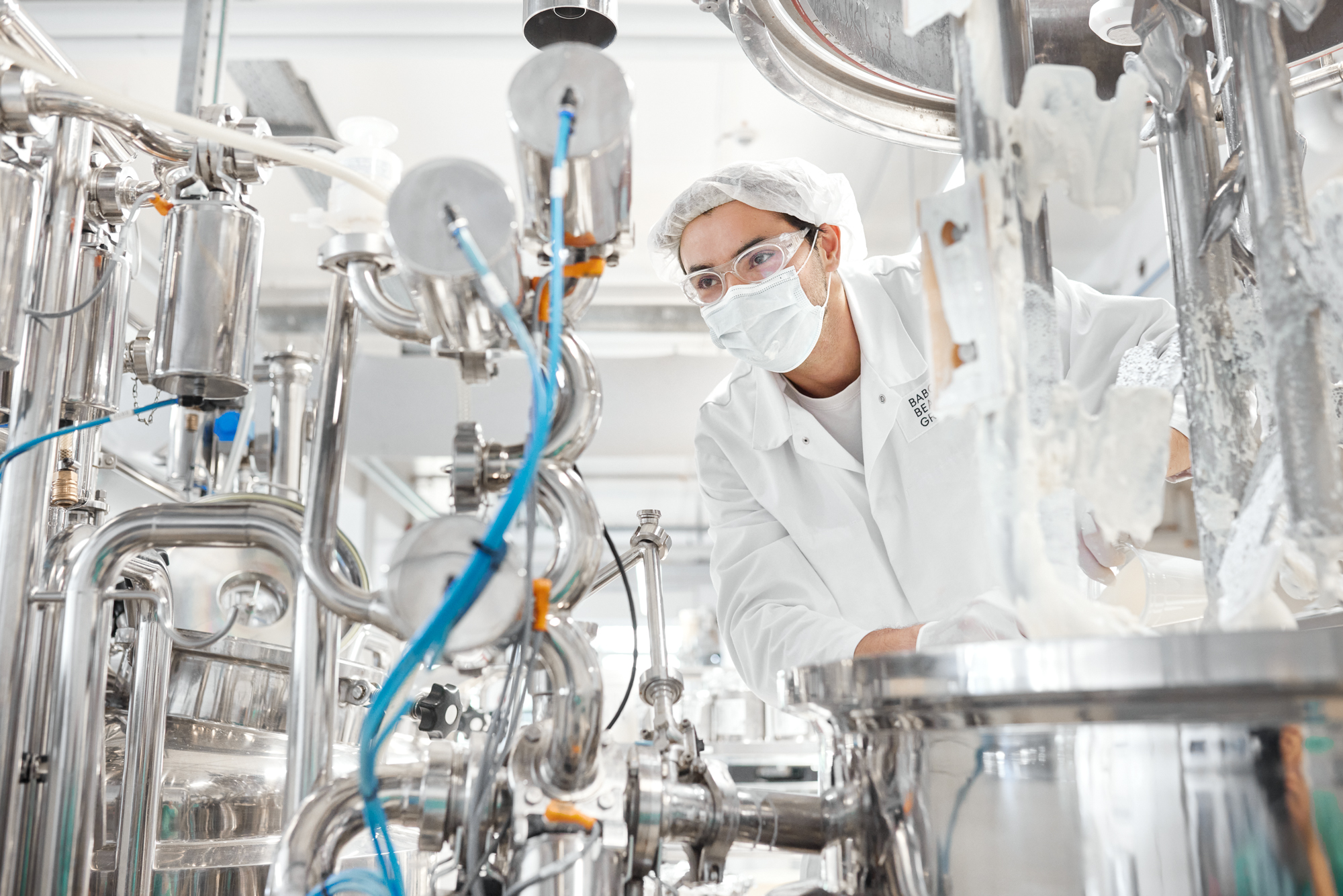 In einer Industrieanlage in Düsseldorf bedient eine Person in Schutzkleidung und Schutzbrille Maschinen, die von Metallrohren und -geräten umgeben sind, und fängt so die Essenz der Unternehmensfotografie für Geschäftsporträts und Reportagen ein. Portrait und Reportage Fotografie für Corporate und Editorial