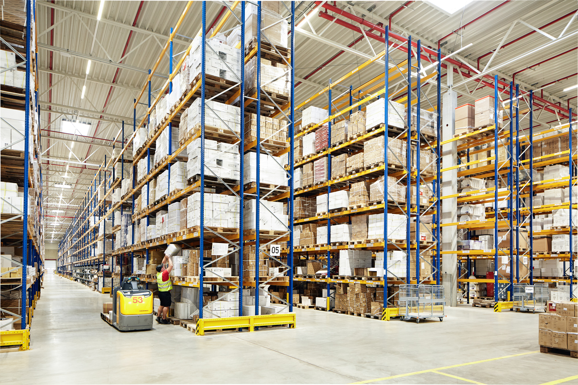 In einem großen Lagerhaus herrscht geschäftiges Treiben. Hohe Regale sind mit Kartons gefüllt, und ein Gabelstapler fährt durch. Es eignet sich ideal für Porträts und Reportagen für Unternehmen und unterstreicht den industriellen Charme der Düsseldorfer Unternehmensfotografie. Portrait und Reportage Fotografie für Corporate und Editorial