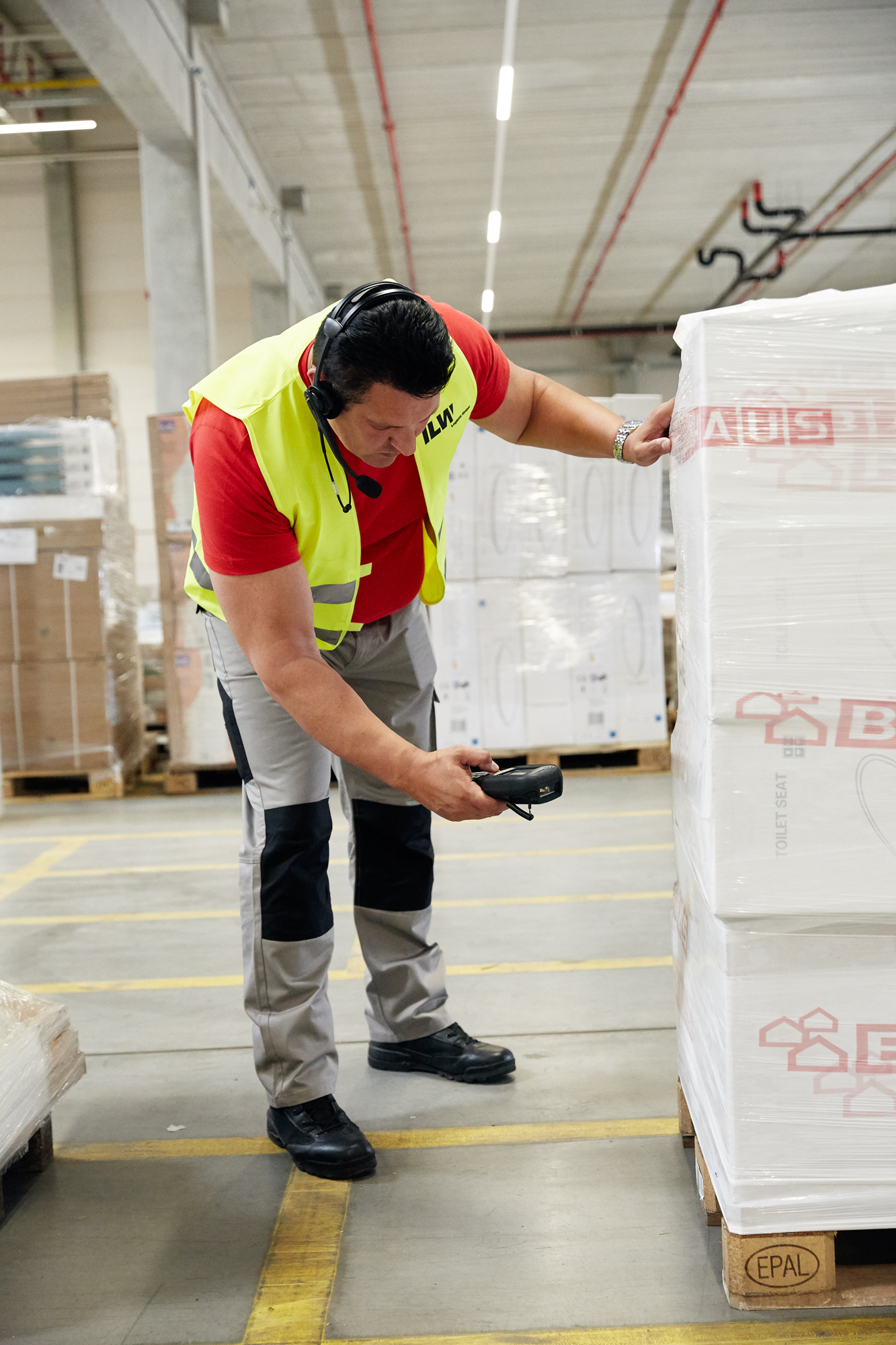 In diesem Beispiel der Unternehmensfotografie Düsseldorf scannt ein Mann in Warnweste und Headset gekonnt mit seinem Handgerät einen Barcode auf einer Palette und veranschaulicht so die dynamische Natur von Reportagen für Unternehmen in geschäftigen Lagerumgebungen. Portrait und Reportage Fotografie für Corporate und Editorial