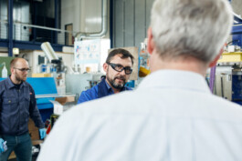 Bei einer Firmenfotosession in Düsseldorf stehen zwei Männer im Vordergrund, während ein dritter Mann mit einer Brille und einer blauen Jacke auf dem Bild steht, während der andere Schutzhandschuhe und ein kariertes Hemd trägt. Um ihn herum sind Maschinen, die sich ideal für Geschäftsporträts und Reportagen eignen. Portrait und Reportage Fotografie für Corporate und Editorial