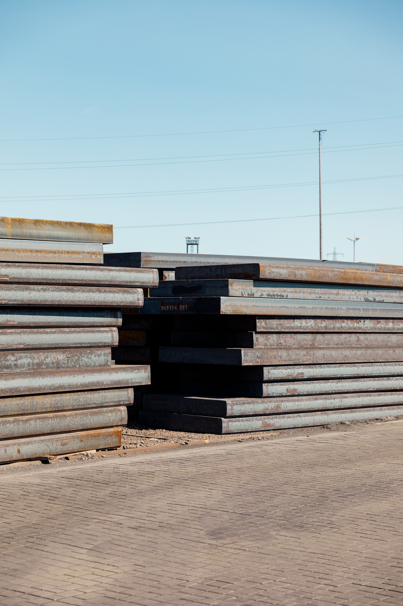 Stapel großer Stahlträger liegen ordentlich auf dem Boden unter einem klaren Himmel und erinnern an Unternehmensfotografien, die Industrielandschaften einfangen. Im Hintergrund ragen hohe Strommasten empor und spiegeln die pulsierende Geschäftsatmosphäre Düsseldorfs wider. Portrait und Reportage Fotografie für Corporate und Editorial