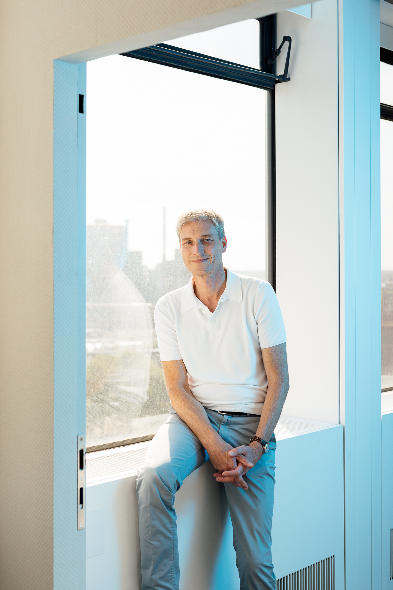 Eine Person mit kurzen Haaren sitzt auf einer Fensterbank. Sie trägt ein weißes Poloshirt und eine helle Hose. In diesem Beispiel der Düsseldorfer Unternehmensfotografie ist der Rahmen perfekt gewählt, durch das Fenster ist eine Stadtlandschaft zu sehen. Portrait und Reportage Fotografie für Corporate und Editorial