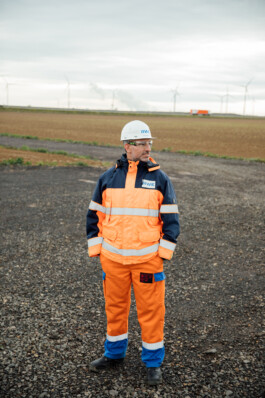Eine Person in leuchtend orangefarbener Arbeitskleidung und mit Helm steht auf einem Kiesfeld, im Hintergrund drehen sich anmutig Windräder – eine perfekte Szene für die Unternehmensfotografie Düsseldorf, die die Essenz des industriellen Fortschritts einfängt. Portrait und Reportage Fotografie für Corporate und Editorial