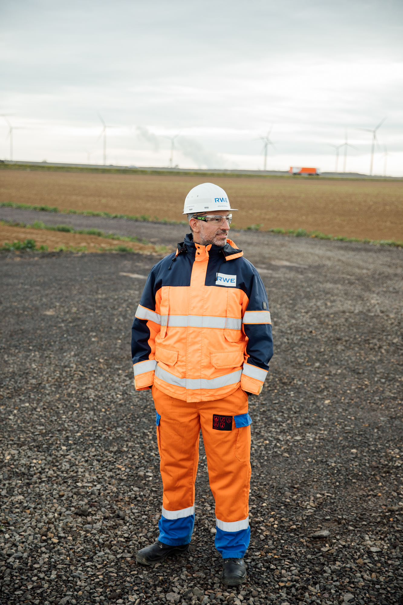 Eine Person in leuchtend orangefarbener Arbeitskleidung und mit Helm steht auf einem Kiesfeld, im Hintergrund drehen sich anmutig Windräder – eine perfekte Szene für die Unternehmensfotografie Düsseldorf, die die Essenz des industriellen Fortschritts einfängt. Portrait und Reportage Fotografie für Corporate und Editorial