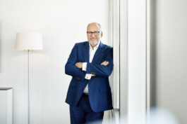 Ein Mann im blauen Anzug steht mit verschränkten Armen neben einer Lampe an einer Wand und strahlt in dem hell erleuchteten Raum Autorität aus. Er beherrscht den Raum mühelos und verkörpert das gelassene Selbstvertrauen, das man von einem Vorstandsmitglied in der dynamischen Geschäftswelt Nordrhein-Westfalens erwarten würde. Portrait und Reportage Fotografie für Corporate und Editorial