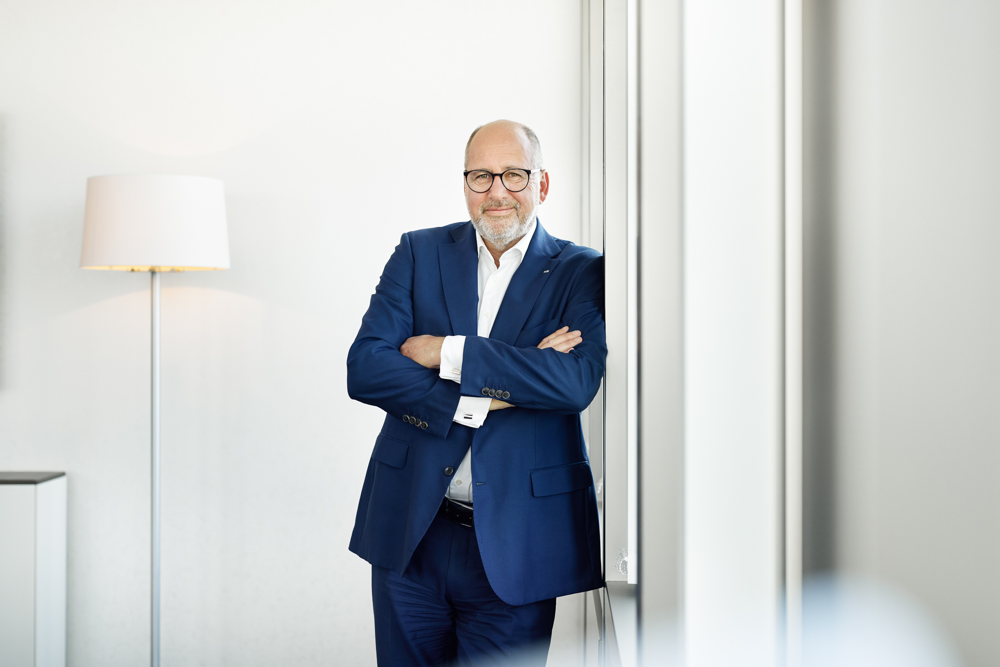 Ein Mann im blauen Anzug steht mit verschränkten Armen neben einer Lampe an einer Wand und strahlt in dem hell erleuchteten Raum Autorität aus. Er beherrscht den Raum mühelos und verkörpert das gelassene Selbstvertrauen, das man von einem Vorstandsmitglied in der dynamischen Geschäftswelt Nordrhein-Westfalens erwarten würde. Portrait und Reportage Fotografie für Corporate und Editorial