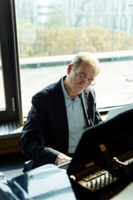 Ein Mann im Anzug spielt im Innenbereich Klavier, eingerahmt von einem großen Fenster mit malerischer Aussicht auf die Umgebung, und fängt die Essenz der Düsseldorfer Unternehmensfotografie für Porträts und Reportagen ein. Portrait und Reportage Fotografie für Corporate und Editorial