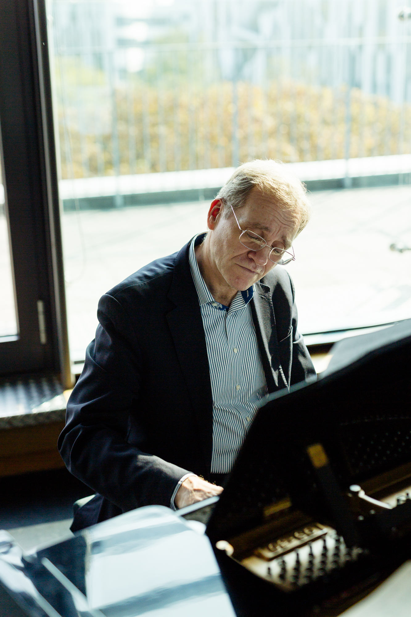 Ein Mann im Anzug spielt im Innenbereich Klavier, eingerahmt von einem großen Fenster mit malerischer Aussicht auf die Umgebung, und fängt die Essenz der Düsseldorfer Unternehmensfotografie für Porträts und Reportagen ein. Portrait und Reportage Fotografie für Corporate und Editorial