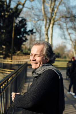 Ein lächelnder Mann mit grauem Haar und schwarzem Schal steht an einem Geländer in einem Park und verströmt mit Bäumen und einem blauen Himmel im Hintergrund einen authentischen Charme. Portrait und Reportage Fotografie für Corporate und Editorial