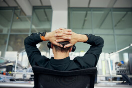 Person im schwarzen Hemd, die mit auf dem Kopf ruhenden Armen an einem Schreibtisch sitzt und auf große Fenster in einer modernen Büroumgebung blickt, was einen authentischen Eindruck der Kontemplation vermittelt. Portrait und Reportage Fotografie für Corporate und Editorial