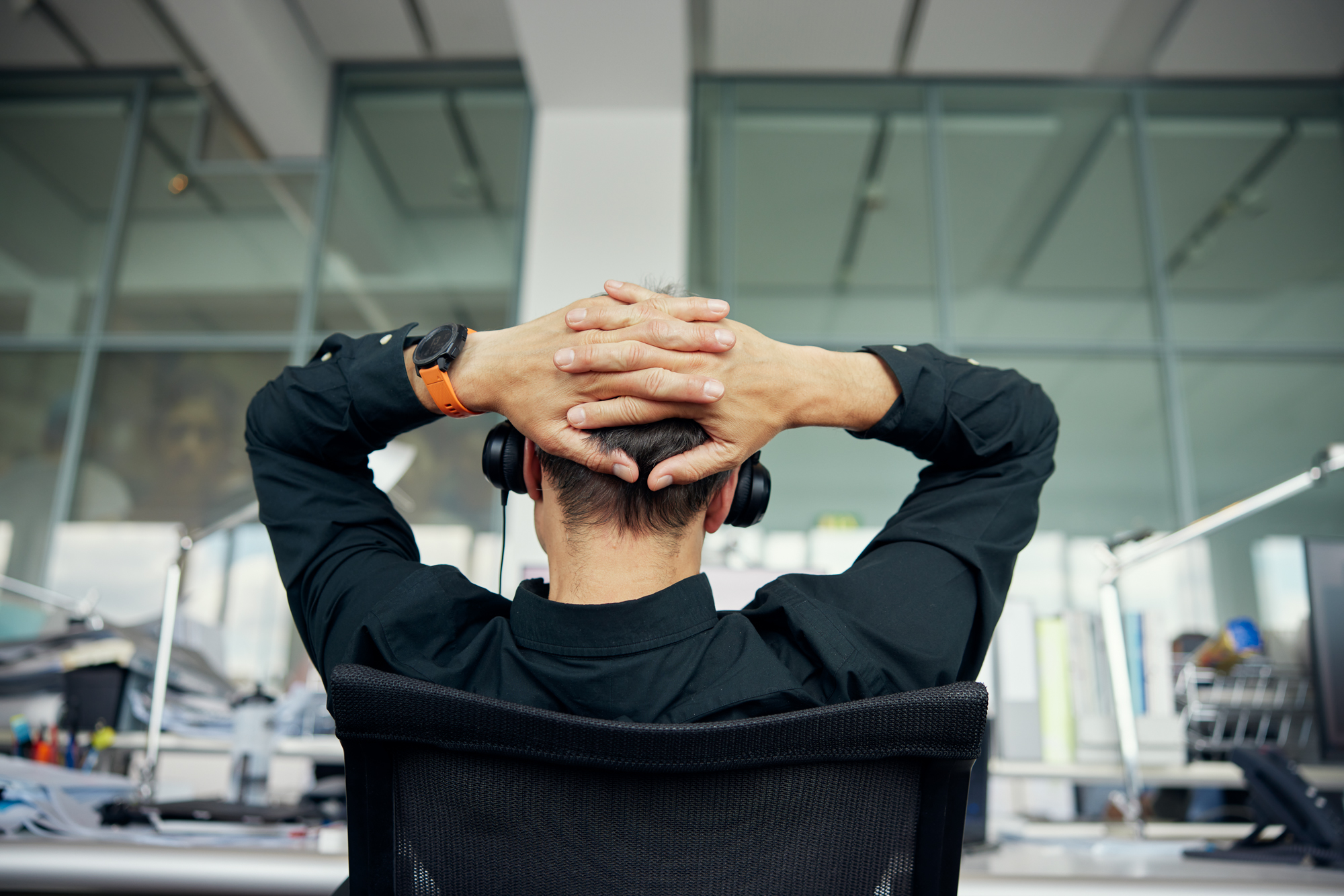 Person im schwarzen Hemd, die mit auf dem Kopf ruhenden Armen an einem Schreibtisch sitzt und auf große Fenster in einer modernen Büroumgebung blickt, was einen authentischen Eindruck der Kontemplation vermittelt. Portrait und Reportage Fotografie für Corporate und Editorial
