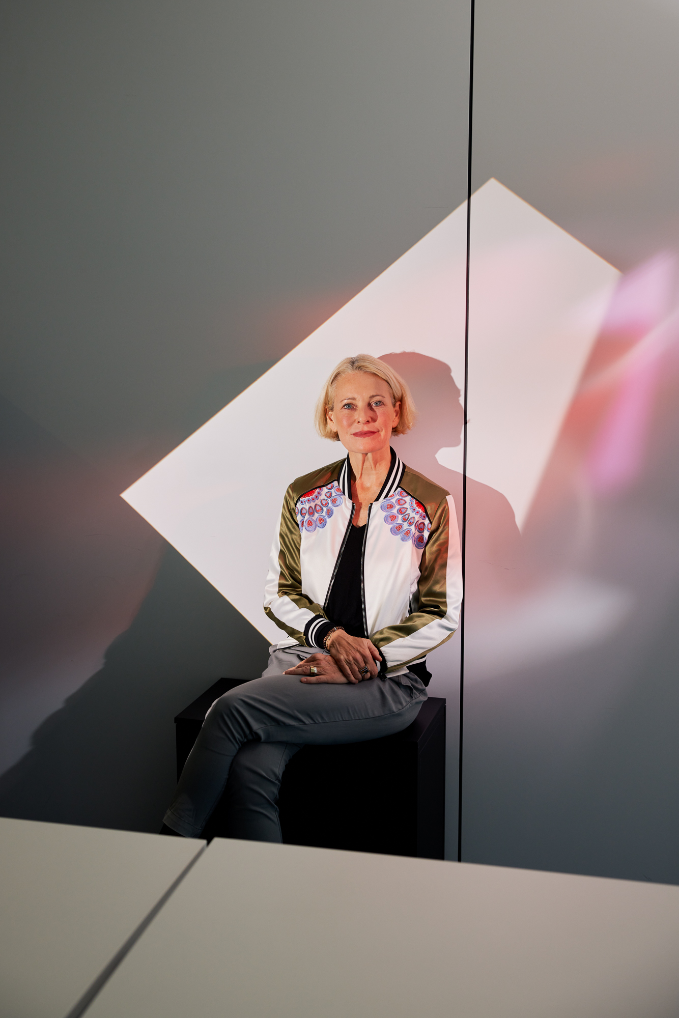 Eine Person mit kurzen Haaren sitzt auf einem Stuhl vor einer geometrisch gemusterten Wand und trägt eine bunte Bomberjacke und graue Hosen – und fängt die Essenz der Corporate Fotografie Düsseldorf ein. Portrait und Reportage Fotografie für Corporate und Editorial