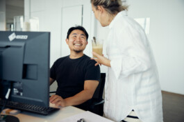 Ein Mann sitzt an seinem Schreibtisch und ist in einen Computerbildschirm vertieft. Als ihm eine Frau neben ihm ein erfrischendes Getränk anbietet, blickt er auf. Dieser spontane Moment fängt die Atmosphäre eines geschäftigen Düsseldorfer Büros ein, wie eine Szene aus einer dieser faszinierenden Reportagen aus dem Alltag. Portrait und Reportage Fotografie für Corporate und Editorial