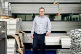 Ein Mann in einem hellblauen Hemd und dunklen Hosen steht in einer Lagerhalle voller gestapelter Metallträger und fängt für ein Employer-Branding-Fotoshooting die Essenz der Unternehmensstärke ein. Portrait und Reportage Fotografie für Corporate und Editorial