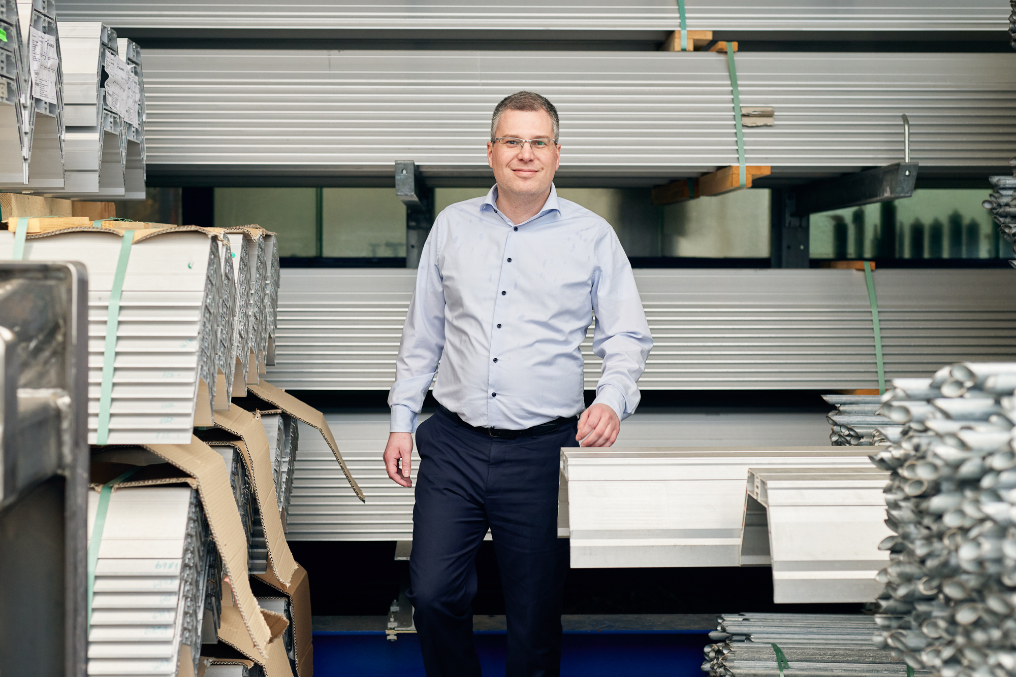Ein Mann in einem hellblauen Hemd und dunklen Hosen steht in einer Lagerhalle voller gestapelter Metallträger und fängt für ein Employer-Branding-Fotoshooting die Essenz der Unternehmensstärke ein. Portrait und Reportage Fotografie für Corporate und Editorial
