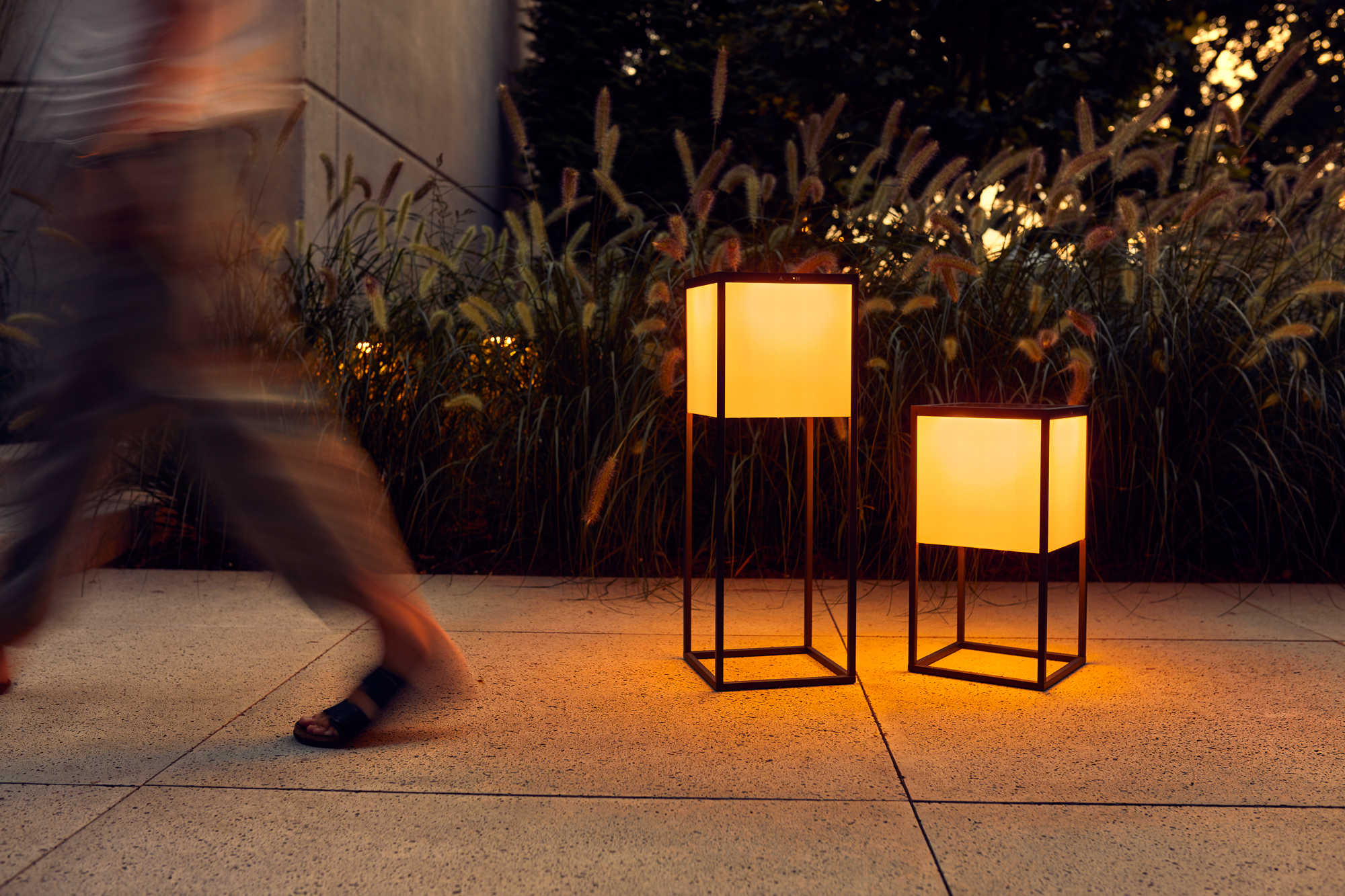 Eine Person geht an zwei beleuchteten quadratischen Laternen auf einem von hohem Gras gesäumten Betonweg vorbei und fängt eine Szene ein, die mühelos als Hintergrund für Firmenporträts dienen könnte. Portrait und Reportage Fotografie für Corporate und Editorial