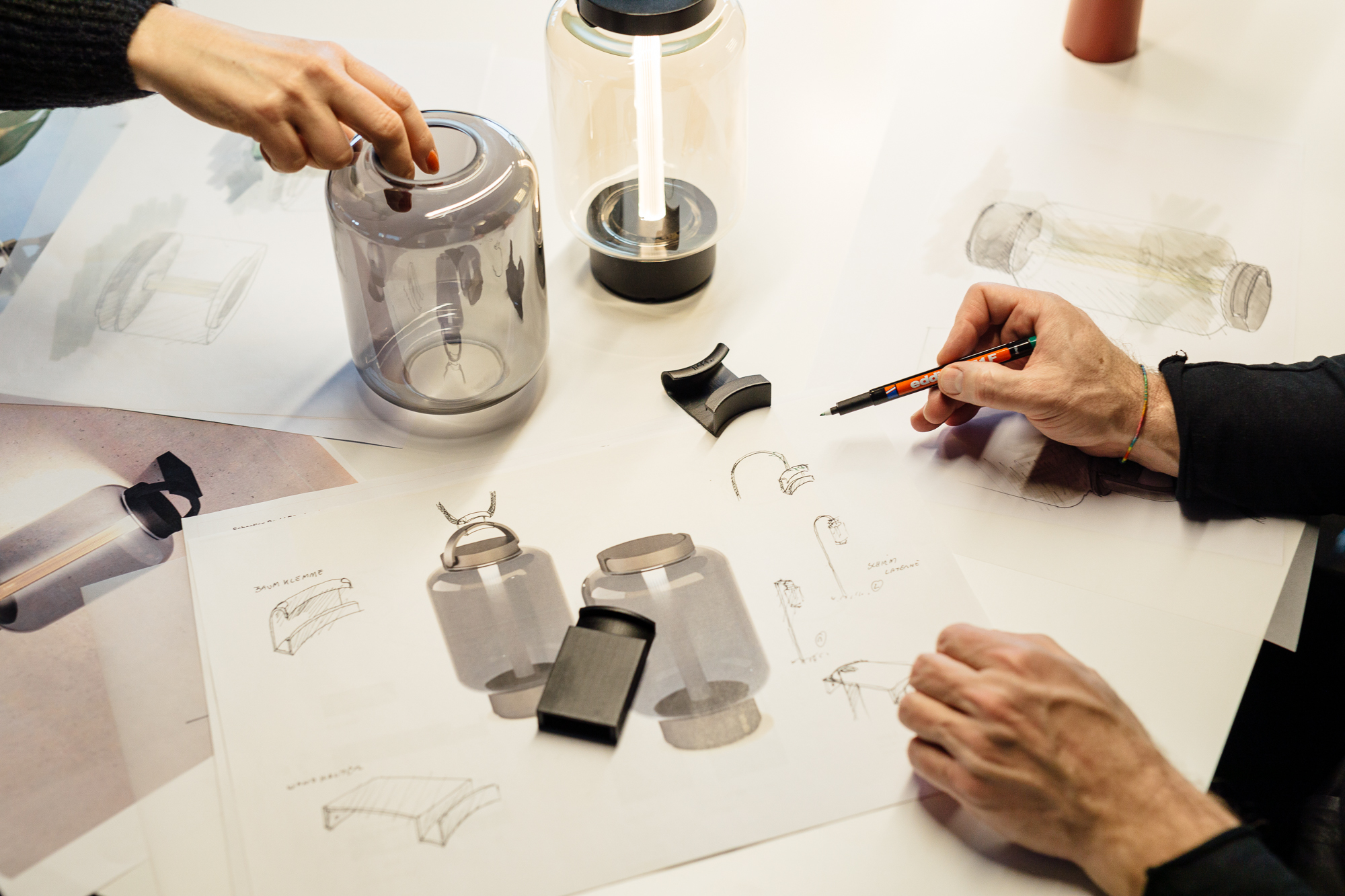 Hände skizzieren und passen die Komponenten eines gefäßähnlichen Objekts auf einem Tisch mit Design-Skizzen an, als würden sie in einem geschäftigen Düsseldorfer Studio den Entwurf für ein innovatives Produkt entwerfen. Portrait und Reportage Fotografie für Corporate und Editorial