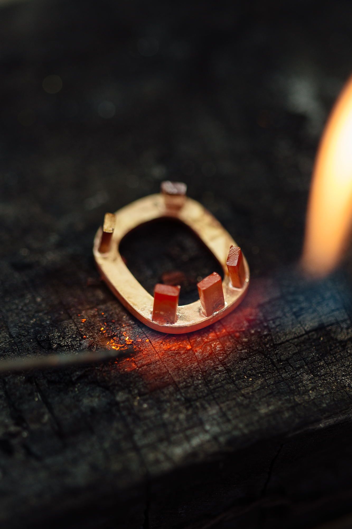 Ein ovales Metallobjekt mit vier Laschen wird erhitzt und wirft in dieser Düsseldorfer Szene einen industriellen Schein auf eine dunkle, verkohlte Oberfläche. Eine kleine Flamme flackert rechts und fesselt das scharfe Auge eines Fotografen, der auf Details und Dynamiken in Unternehmen spezialisiert ist. Portrait und Reportage Fotografie für Corporate und Editorial