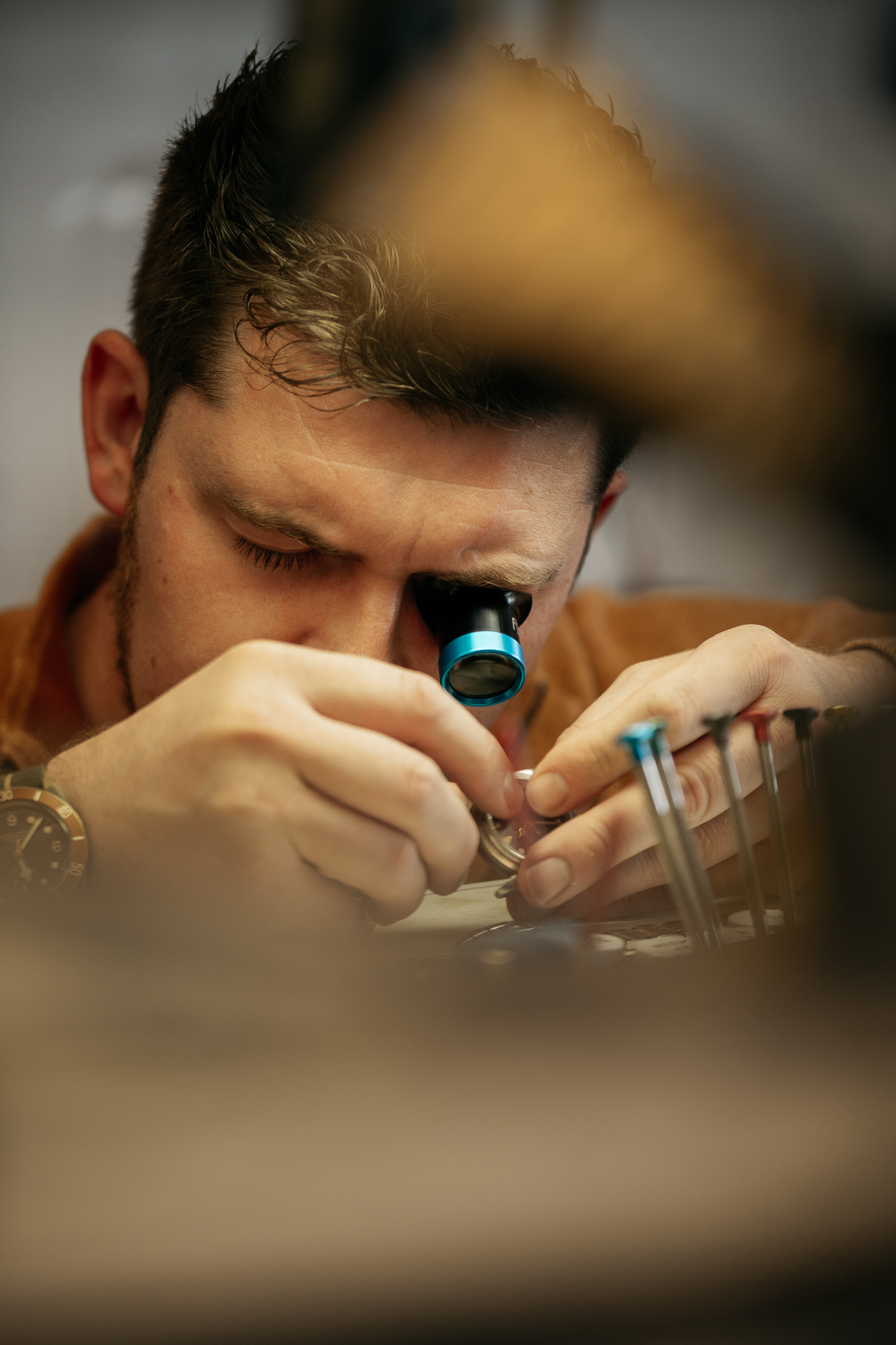 Mit höchster Konzentration bearbeitet eine Person mithilfe einer Lupe und Präzisionswerkzeugen ein kleines mechanisches Objekt. Diese Szene könnte auch Teil einer Employer-Branding-Reportage sein und zeigt die Hingabe und Handwerkskunst, die in jedem Detail steckt. Portrait und Reportage Fotografie für Corporate und Editorial