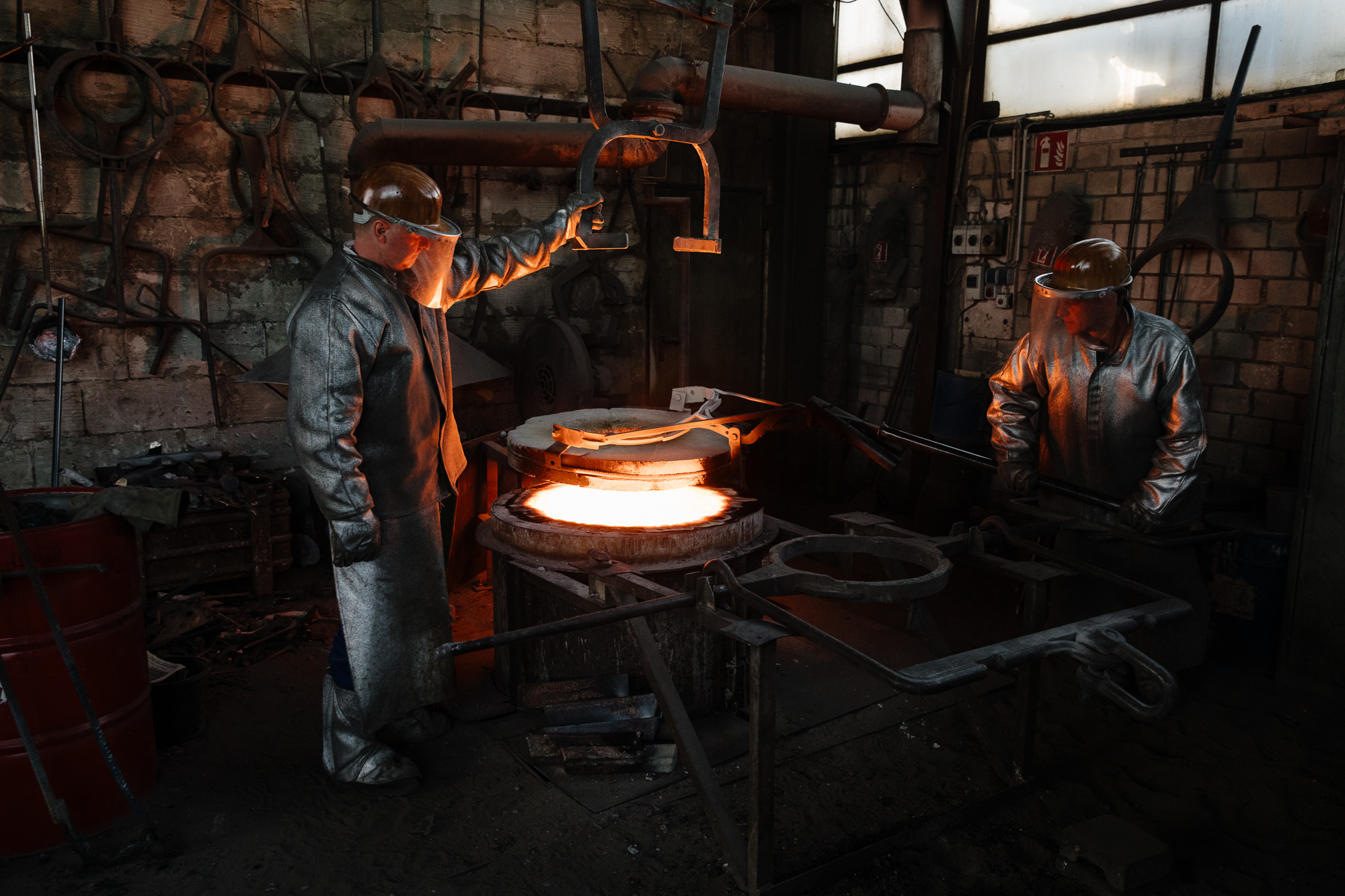Zwei Arbeiter in Schutzanzügen und Helmen bedienen Geräte neben einem glühenden Ofen in einer Industrieanlage und fangen mit ihrer Hingabe die Essenz einer Reportage ein – eine Szene, die von einem erfahrenen Fotografen mit Porträt-Expertise leicht verewigt werden könnte. Portrait und Reportage Fotografie für Corporate und Editorial