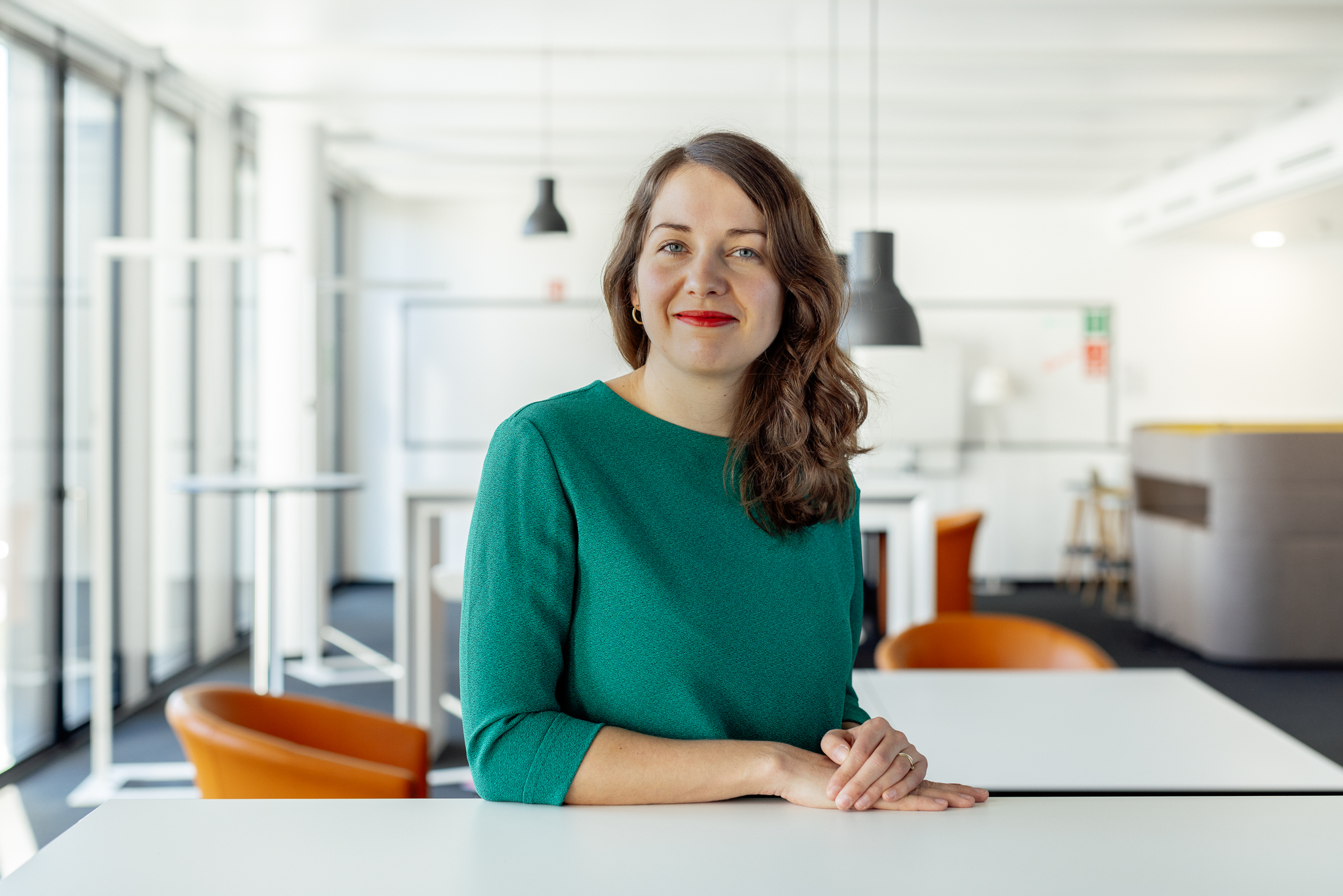 Eine Frau in einem grünen Oberteil lächelt und verkörpert ein perfektes Unternehmensporträt, während sie an einem weißen Tisch in einem hellen, modernen Büroraum mit orangefarbenen Stühlen und großen Fenstern sitzt. Portrait und Reportage Fotografie für Corporate und Editorial