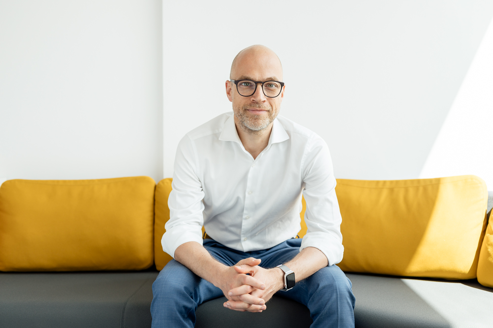 Ein kahlköpfiger Mann mit Brille sitzt auf einem grau-gelben Sofa. Er trägt ein weißes Hemd und eine blaue Hose, die Hände sind gefaltet, und verkörpert das ruhige und doch konzentrierte Auftreten, das oft auf Firmenporträts zu sehen ist. Portrait und Reportage Fotografie für Corporate und Editorial