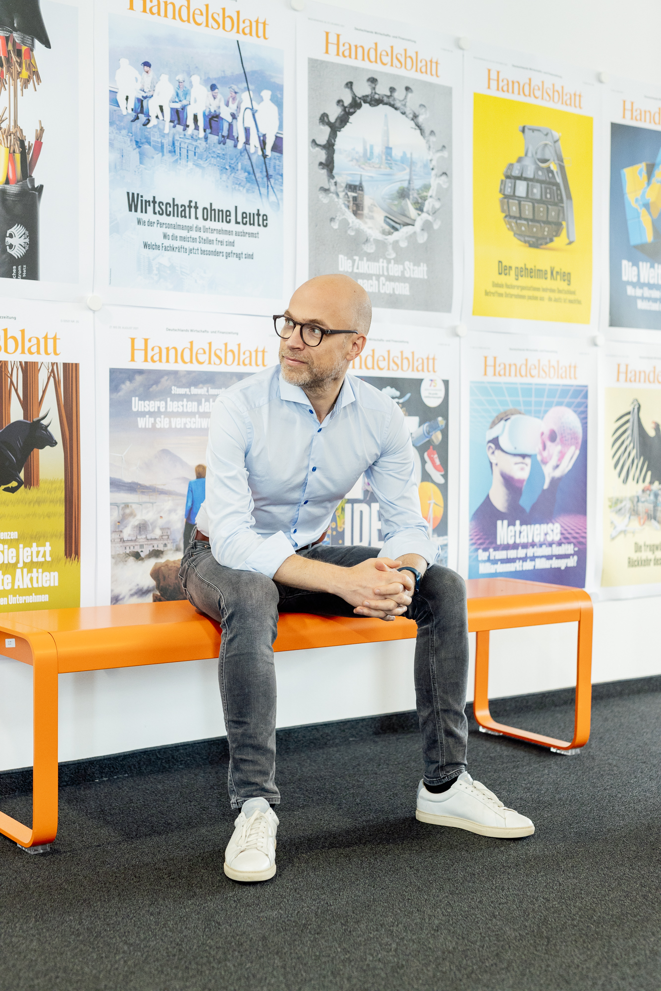 Auf einer orangefarbenen Bank sitzt eine Person in hellblauem Hemd und Jeans und betrachtet die bunten Zeitungscover an der Wand. Es ist eine Szene, die Employer-Branding-Experten und Storytelling-Enthusiasten gleichermaßen anspricht, vielleicht direkt aus Düsseldorfs Kreativzentren. Portrait und Reportage Fotografie für Corporate und Editorial