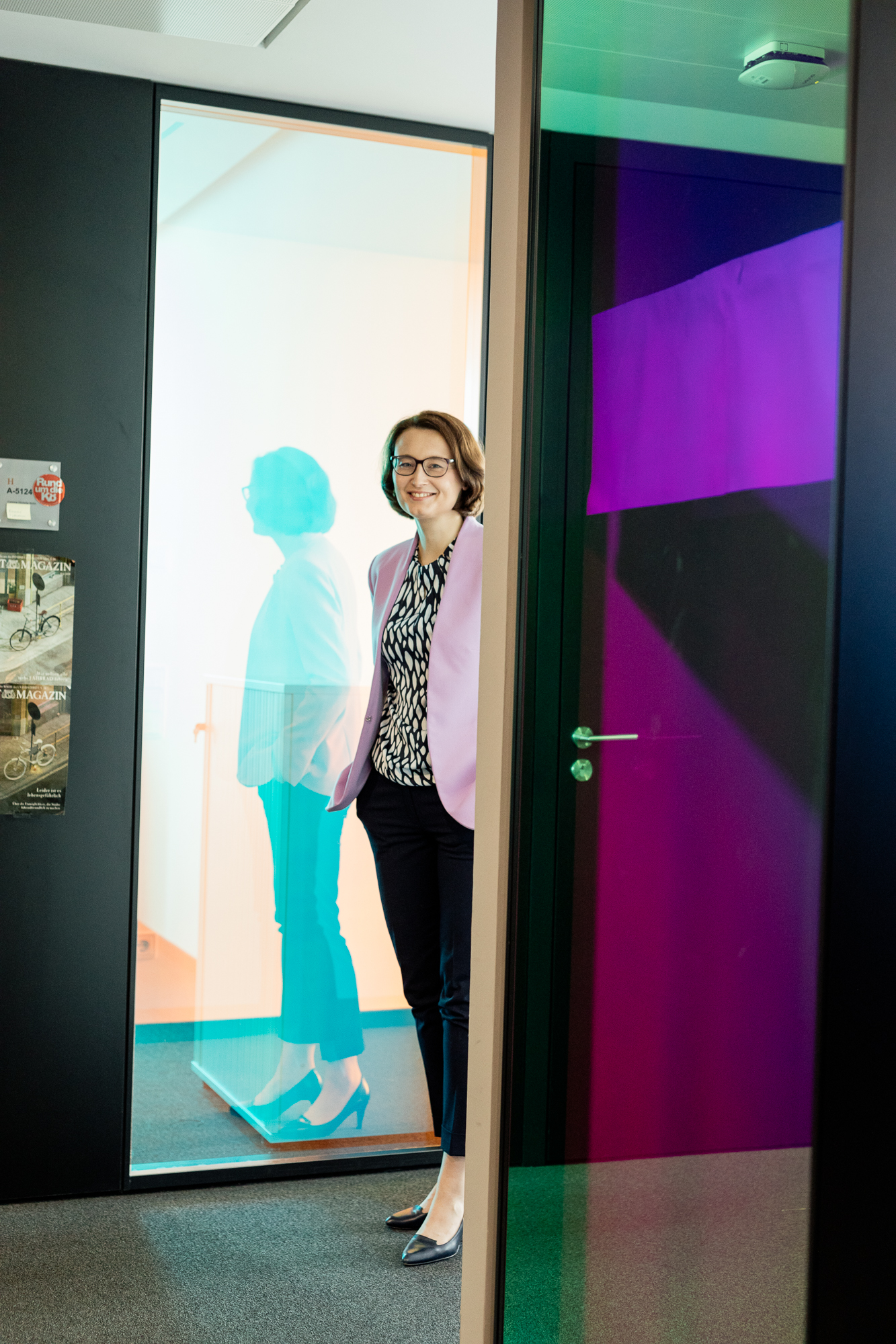 In einem lebhaften Flur steht eine lächelnde Person in einem lila Blazer und einer schwarzen Hose. Bunte Glasplatten werfen strahlende Reflexionen und fangen die Szene ein, wie ein Fotograf, der geschäftliche Momente festhält. Poster schmücken die Wände und verleihen dieser dynamischen Umgebung eine kreative Note. Portrait und Reportage Fotografie für Corporate und Editorial