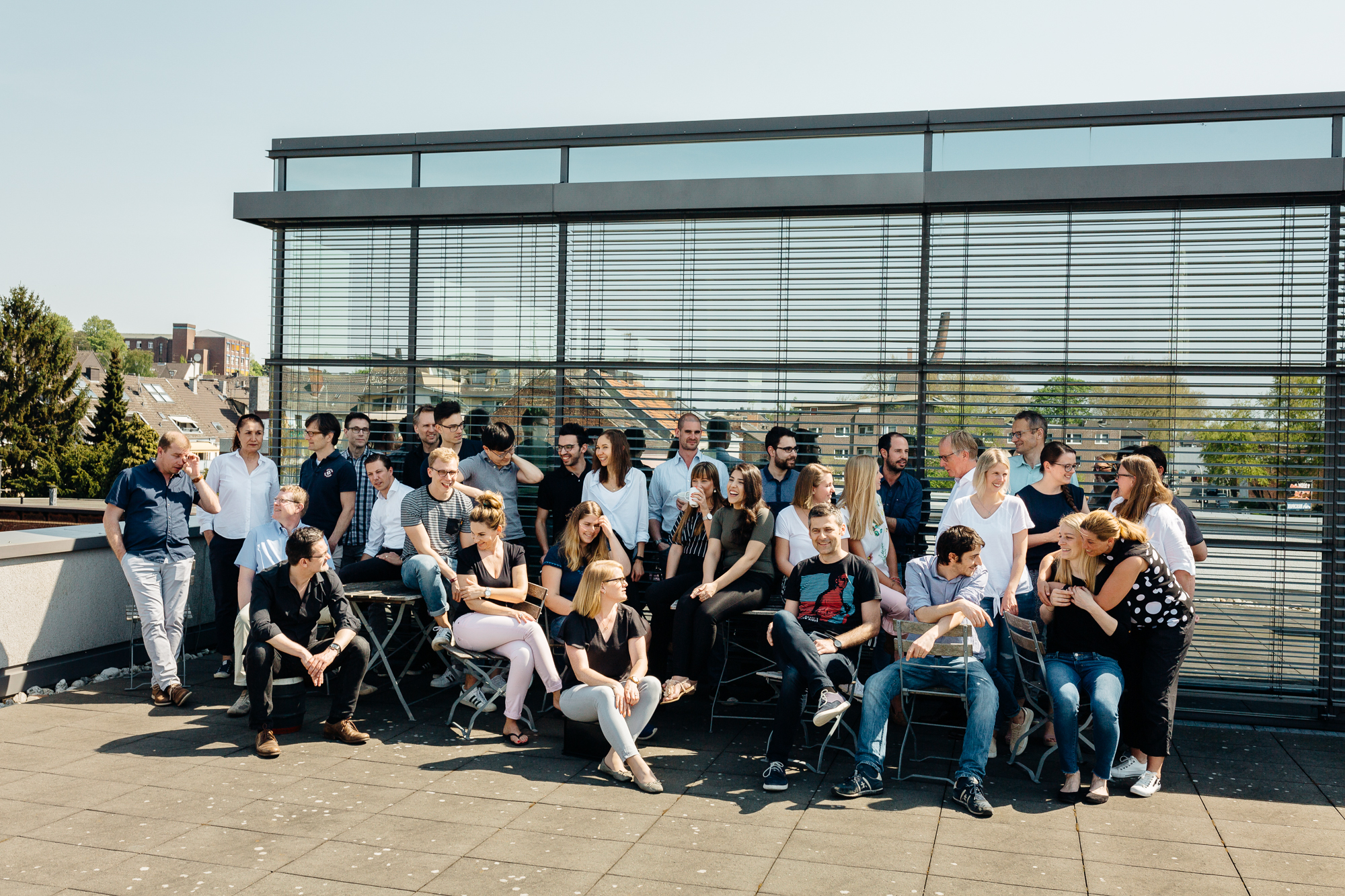 Eine Gruppe von Menschen posiert gemeinsam auf einer sonnigen Dachterrasse in Düsseldorf, eingerahmt von großen Fenstern. Einige sitzen, andere stehen und schaffen so eine einladende Szene, die den Geist des modernen Employer Branding durch die Linse eines talentierten Fotografen einfängt. Portrait und Reportage Fotografie für Corporate und Editorial