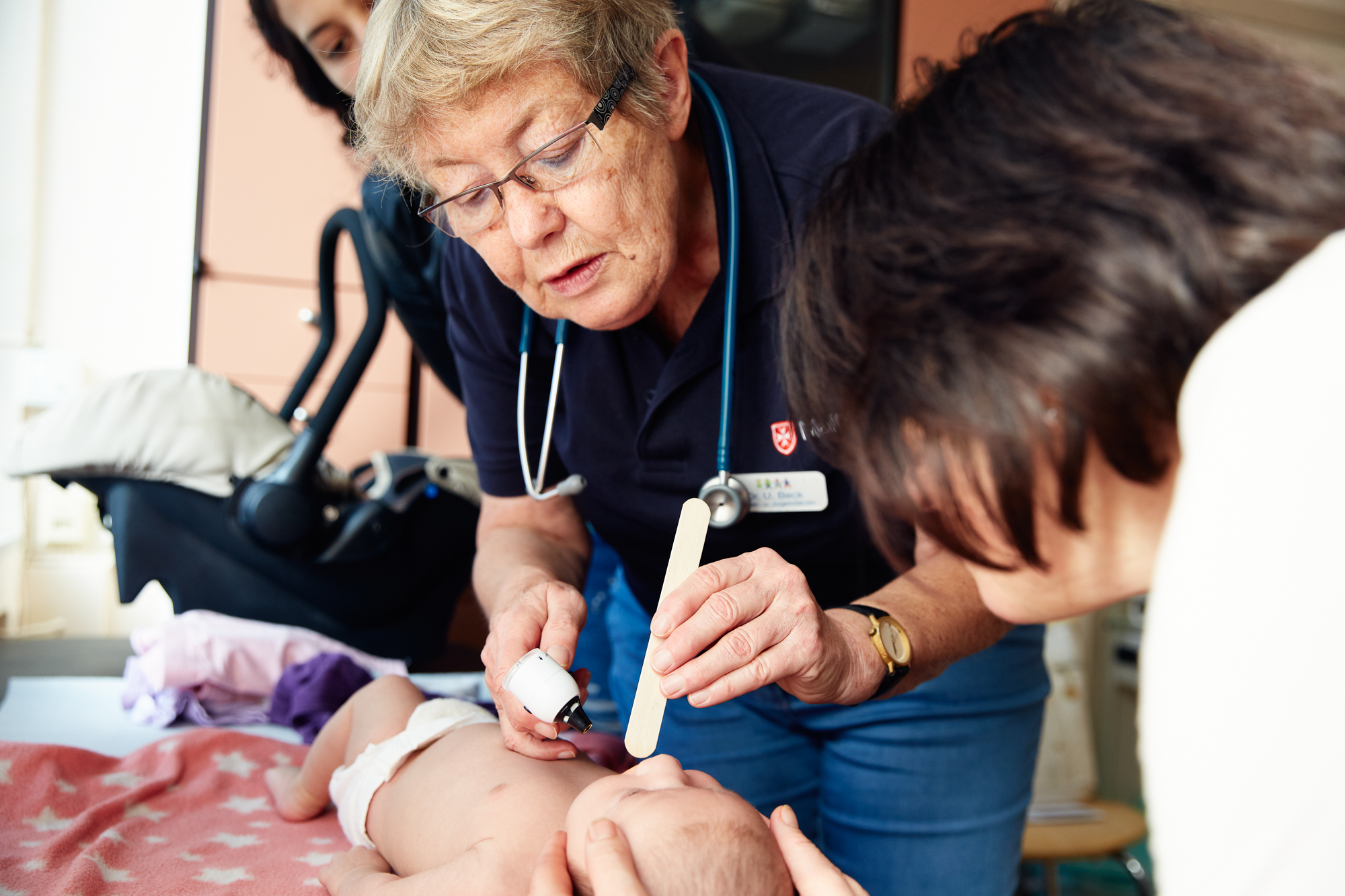 Eine medizinische Fachkraft in Düsseldorf untersucht den Bauch eines Babys sorgfältig mit einem medizinischen Instrument, während ein anderer Erwachsener in der Nähe zusieht und den Moment festhält, wie ein erfahrener Fotograf, der eine aufschlussreiche Reportage erstellt. Portrait und Reportage Fotografie für Corporate und Editorial