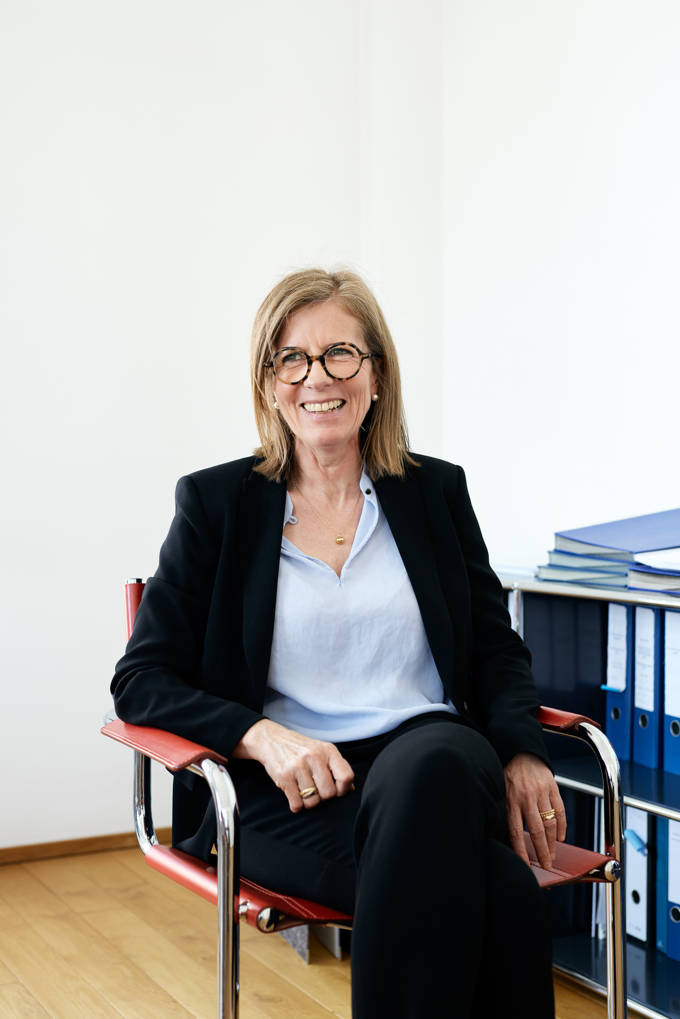 Auf einem Bürostuhl sitzt eine Person mit Brille, schwarzem Blazer und weißem Hemd und lächelt selbstbewusst mit den Händen auf den Armlehnen. Das Firmenambiente wird durch die helle Wand und die gestapelten blauen Ordner hervorgehoben, die vielleicht einen Moment für einen Düsseldorfer Fotografen festhalten. Portrait und Reportage Fotografie für Corporate und Editorial