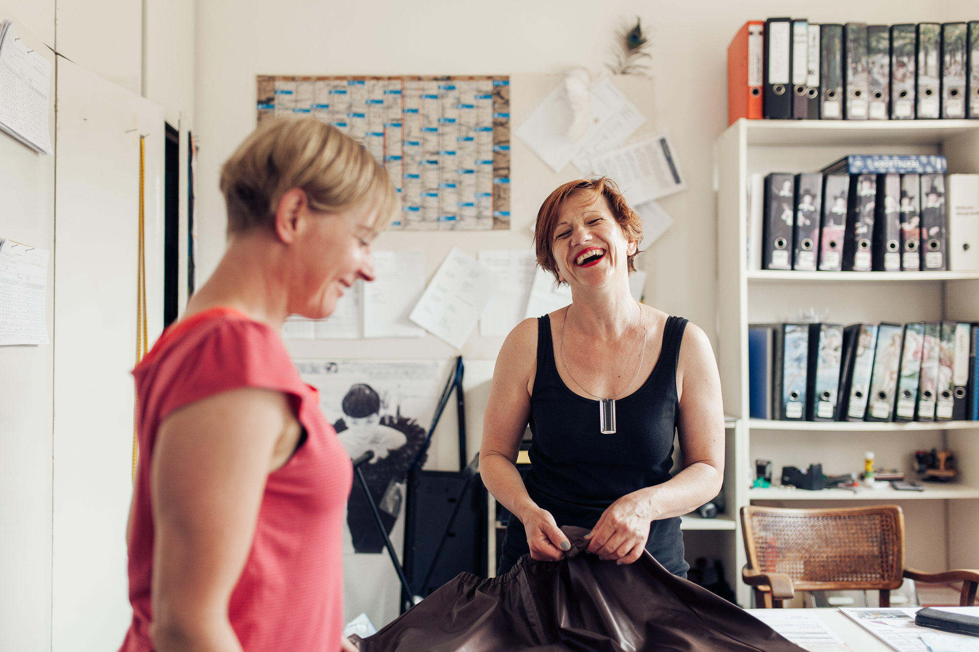 In einem geschäftigen Büro voller Ordner und Papiere werden zwei Frauen in einem spontanen Moment fotografiert. Eine lächelt breit und hält einen Stoff, während die andere zusieht. Diese lebhafte Szene ruft die Essenz des Unternehmenslebens durch die Reportagenlinse eines scharfsinnigen Fotografen hervor. Portrait und Reportage Fotografie für Corporate und Editorial