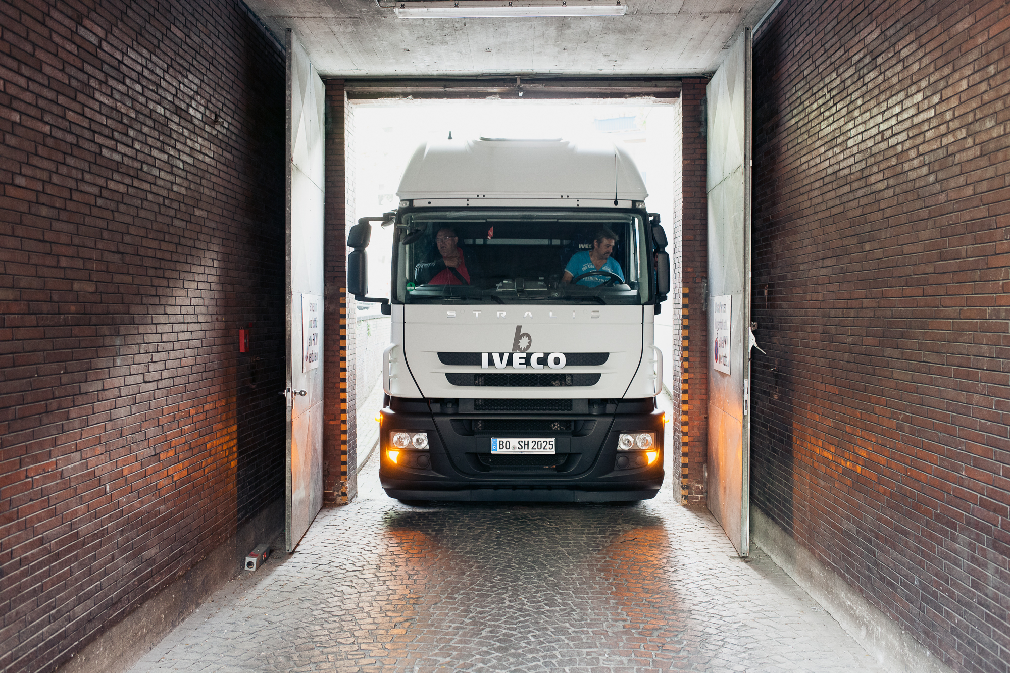 Ein weißer Iveco-Truck navigiert gekonnt durch eine schmale, von Backsteinmauern umgebene Passage und gleicht einem Industrieporträt. Durch die Windschutzscheibe sind zwei Personen in der Kabine zu sehen, die konzentrierte Blicke austauschen. Portrait und Reportage Fotografie für Corporate und Editorial
