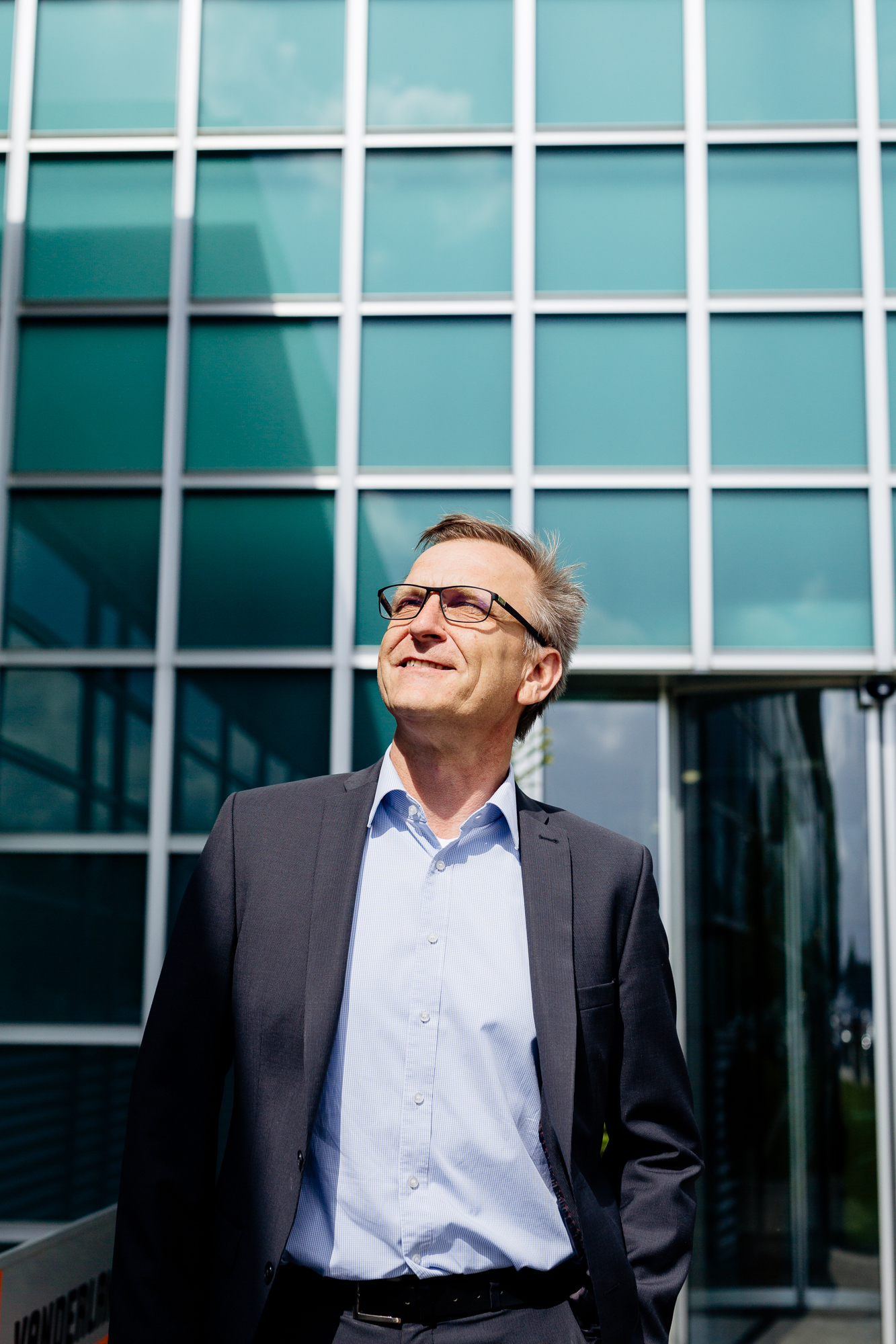 Ein Mann im Anzug steht vor einem modernen Glasgebäude, blickt mit einem leichten Lächeln nach oben und verkörpert das Selbstbewusstsein eines Unternehmens. Portrait und Reportage Fotografie für Corporate und Editorial