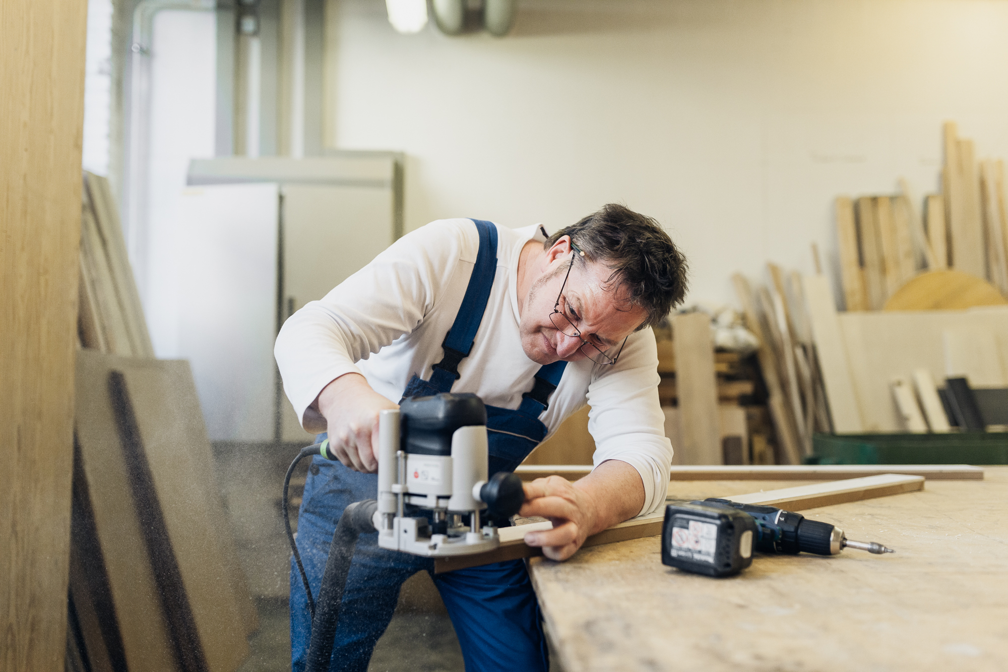 Ein Mann im Overall schwingt gekonnt ein Elektrowerkzeug auf einem Holzbrett in einer geschäftigen Werkstatt, umgeben von einer Reihe von Holzbearbeitungsmaterialien. Diese Szene könnte von jedem begeisterten Fotografen festgehalten werden, der die Handwerkskunst des Handwerkers für Arbeitgebermarkenzwecke hervorheben möchte. Portrait und Reportage Fotografie für Corporate und Editorial