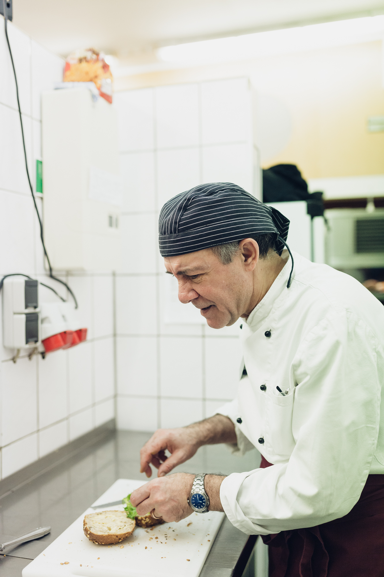 In einer geschäftigen Düsseldorfer Küche schneidet ein Koch in weißem Kittel und gestreifter Mütze gekonnt Zutaten auf einem Schneidebrett. Die Szene fängt die Essenz kulinarischer Kunst ein und ist perfekt für jeden aufstrebenden Fotografen, der das Employer Branding in der kulinarischen Welt hervorheben möchte. Portrait und Reportage Fotografie für Corporate und Editorial