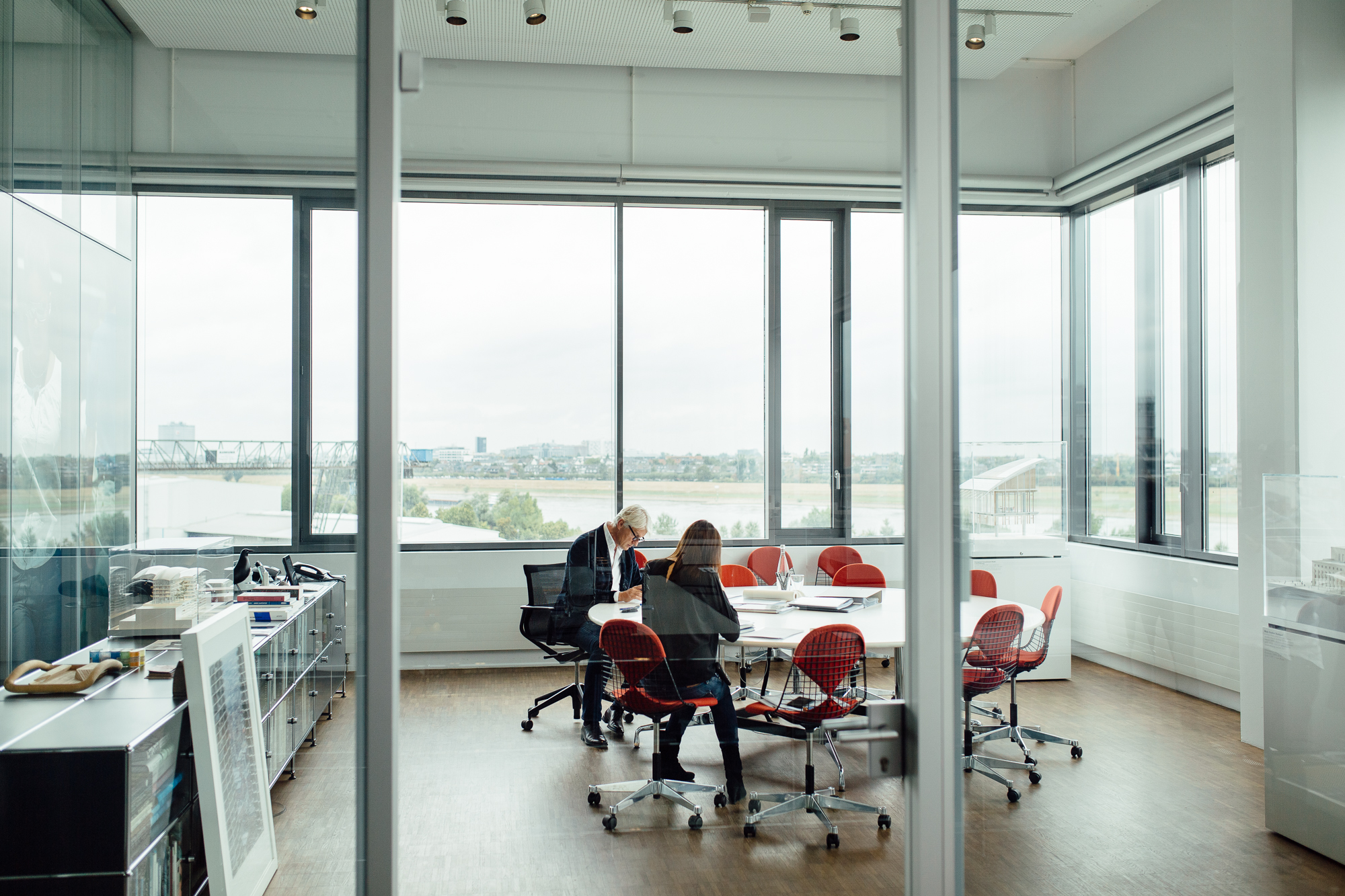 Zwei Personen sitzen an einem runden Tisch in einem Büro mit Glaswänden und roten Stühlen und prüfen Dokumente. Große Fenster bieten einen atemberaubenden Blick auf den Fluss und die Skyline von Düsseldorf und bilden eine perfekte Kulisse für diese Unternehmensumgebung, die an die Komposition eines erfahrenen Fotografen erinnert. Portrait und Reportage Fotografie für Corporate und Editorial