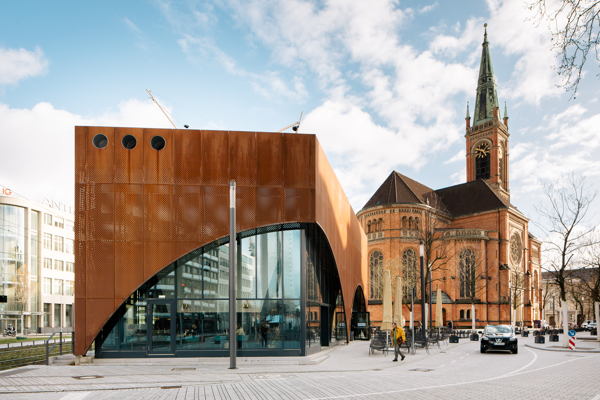 Ein modernes Gebäude mit rostfarbener Fassade steht neben einer historischen Kirche, deren hoher Turm in den teilweise bewölkten Himmel ragt. Diese in lebendigen Details festgehaltene Szene des Alltags wurde von einem talentierten Fotografen im Rahmen seiner Architekturreportage verewigt. Portrait und Reportage Fotografie für Corporate und Editorial