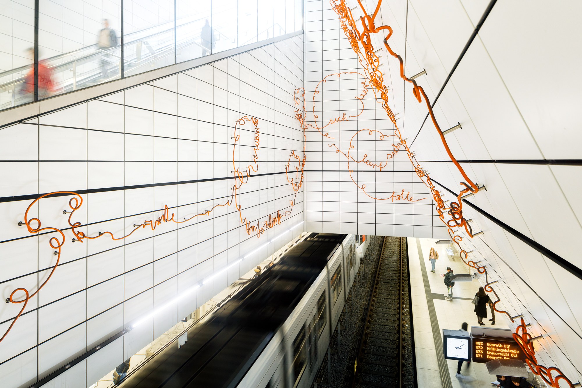 Eine moderne U-Bahnstation mit kunstvollen orangefarbenen Linien auf weiß gefliesten Wänden spiegelt die Essenz der urbanen Eleganz Düsseldorfs wider. Unten wartet ein Zug, während Menschen den Bahnsteig überqueren, eingerahmt wie eine Firmenszene in der Linse eines Fotografen, unter einer beleuchteten Informationstafel. Portrait und Reportage Fotografie für Corporate und Editorial