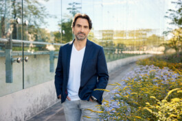 Ein Mann mit dunklem Haar und Bart steht im Freien und sieht aus wie ein Reportagefotograf aus Düsseldorf. Er trägt einen marineblauen Blazer und ein weißes Hemd auf einem mit Glasgeländern und gelben Blumen geschmückten Weg. Portrait und Reportage Fotografie für Corporate und Editorial